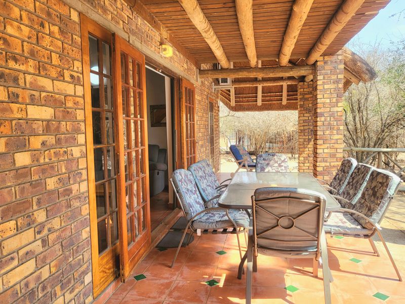 A covered outdoor patio featuring exposed brickwork, a rustic timber beam ceiling with thatch, and terracotta tiled flooring, providing direct access to the interior.