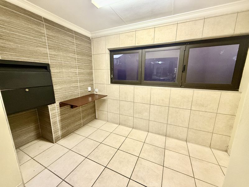 A clean, tiled patio area featuring a built-in braai, wall-mounted shelf, and large aluminium-framed windows, offering a functional space for outdoor entertaining.