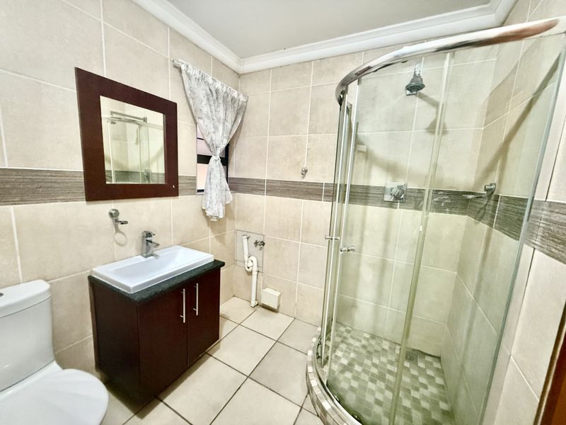 A functional bathroom featuring a corner shower enclosure, vanity basin, and toilet, finished with neutral wall and floor tiling.