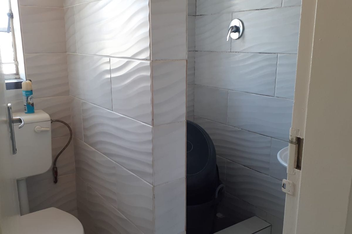 A compact bathroom featuring textured white wall tiles, a standard toilet, and a shower area with a wall-mounted mixer.