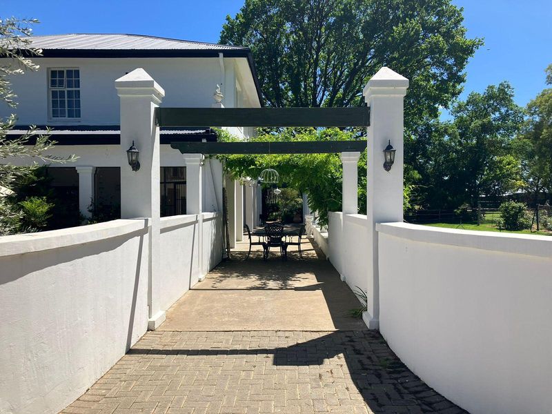 A charming outdoor patio area featuring a pergola, paved walkway, and white masonry pillars, providing a shaded transition space alongside the main farmhouse structure.