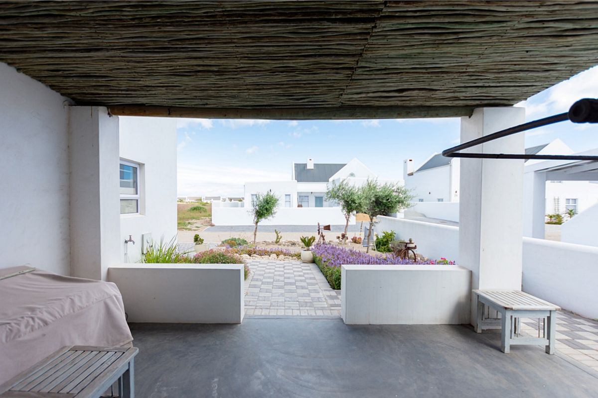 A covered patio featuring a rustic thatched roof and concrete flooring, overlooking a landscaped garden and paved pathway.