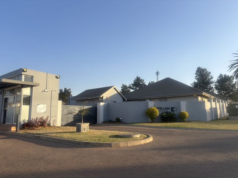 Front entrance view of the townhouse complex featuring a security gatehouse, paved driveway, and perimeter wall with security fencing.