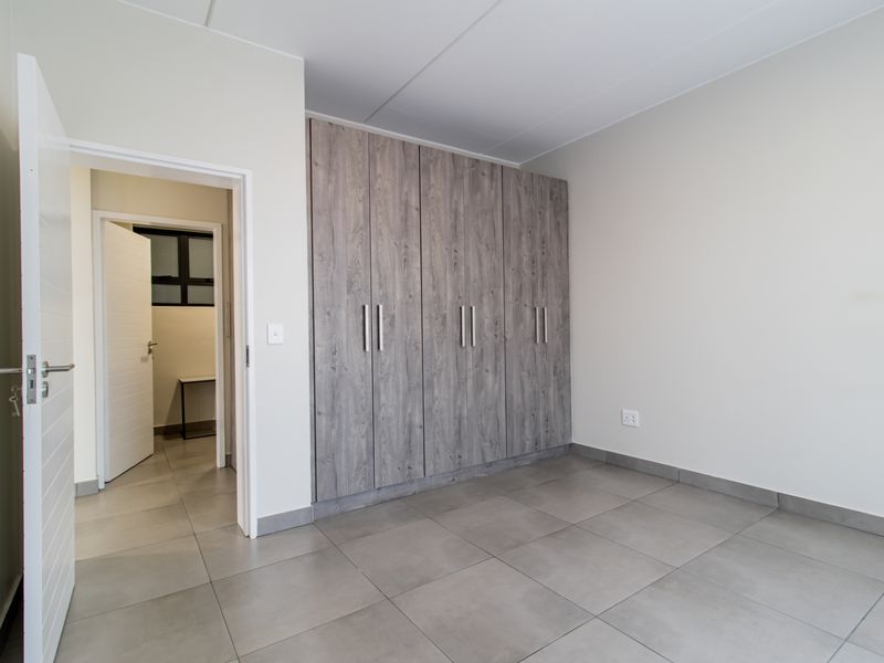 A clean, empty bedroom featuring modern grey tiled flooring and built-in wooden cupboards, with an adjacent hallway visible through the doorway.