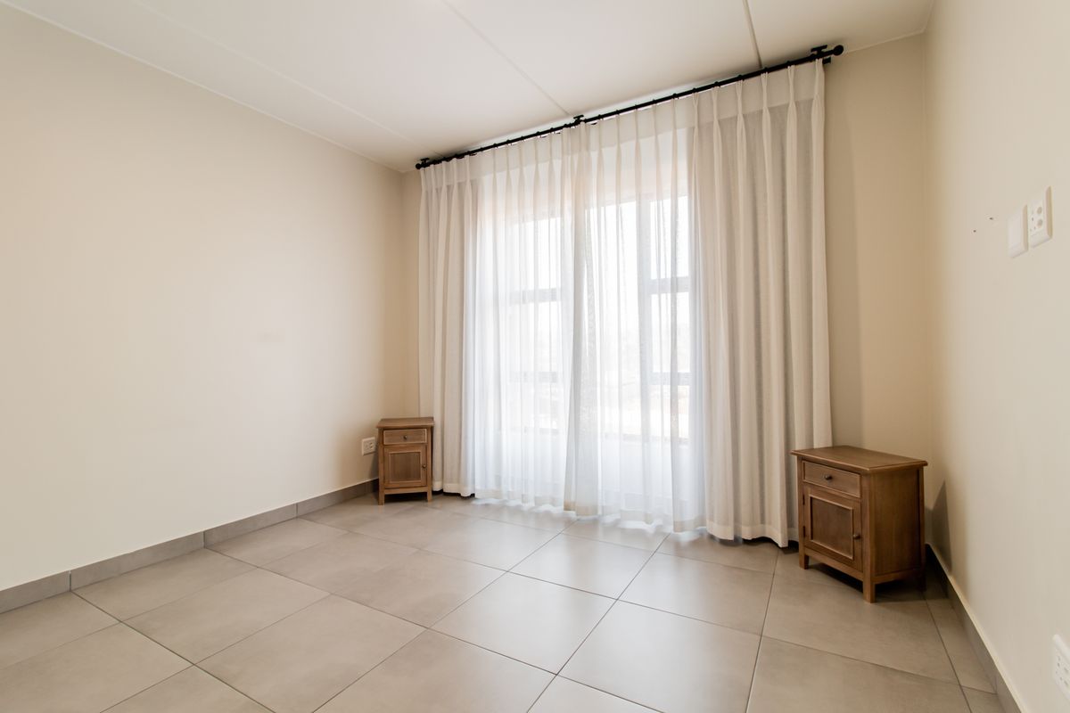 A bright, neutral-toned bedroom featuring large windows with floor-to-ceiling curtains and modern tiled flooring, offering a clean and versatile living space.