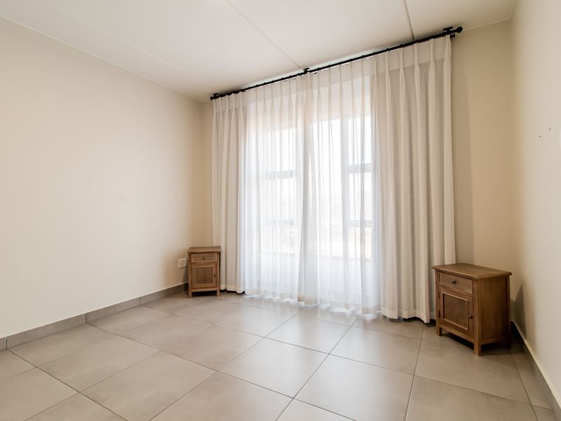 A bright, neutral-toned bedroom featuring large windows with floor-to-ceiling curtains and modern tiled flooring, offering a clean and versatile living space.