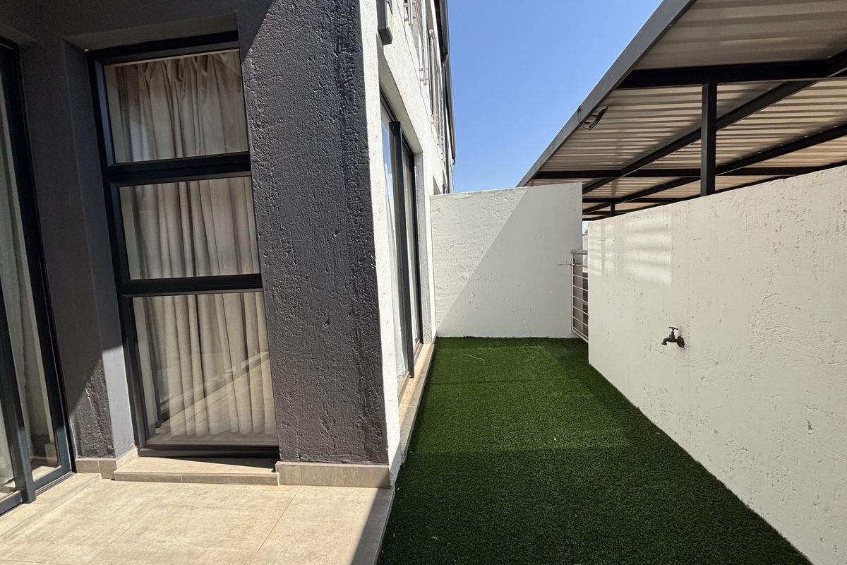 A neat rear exterior area featuring a tiled patio, artificial grass lawn, and a covered walkway along the side of the townhouse.