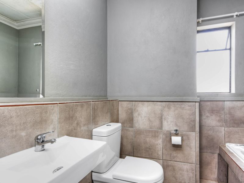 A functional bathroom featuring a wall-mounted basin, dual-flush toilet, and a built-in bathtub with neutral wall tiling and a large mirror.