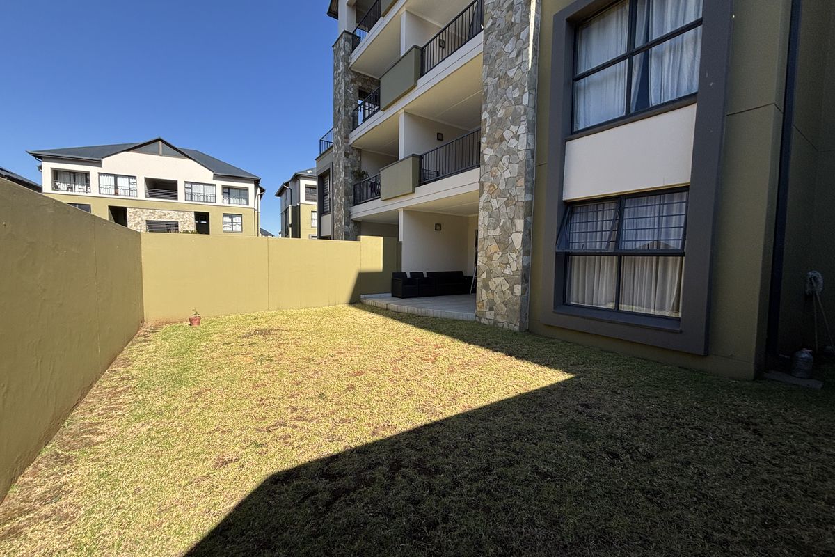 A private rear garden area featuring a paved patio, lawn, and stone-clad building facade with multi-level balconies.