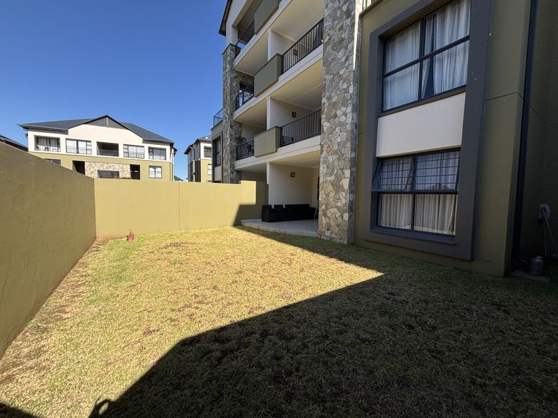 A private rear garden area featuring a paved patio, lawn, and stone-clad building facade with multi-level balconies.