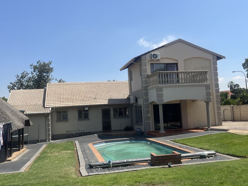 Rear view of a two-story house featuring a swimming pool, paved patio area, upper-level balcony, and a detached thatched lapa in the garden.