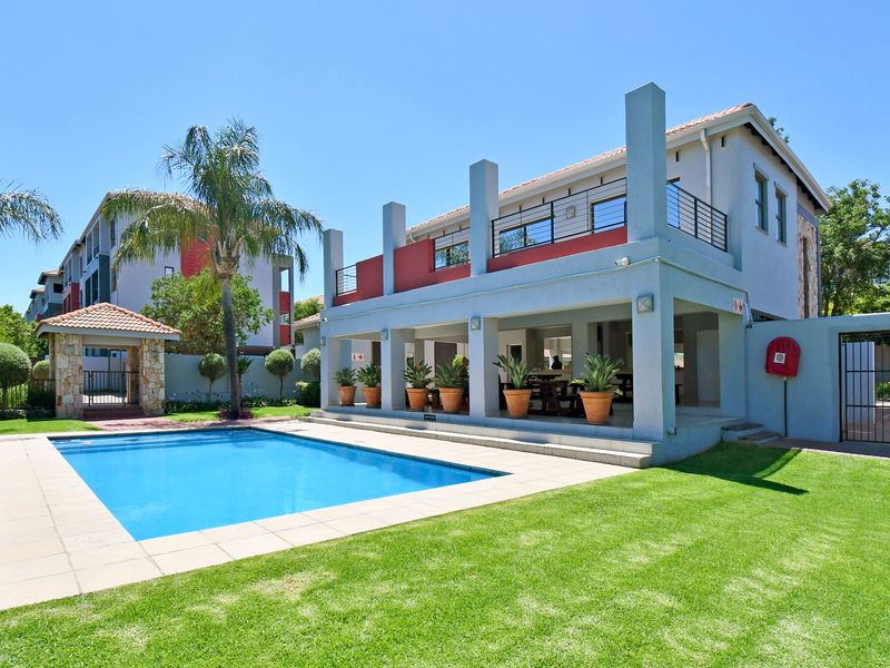 A well-maintained communal pool area featuring a swimming pool, tiled surround, landscaped garden, and a covered patio with an upper-level balcony.