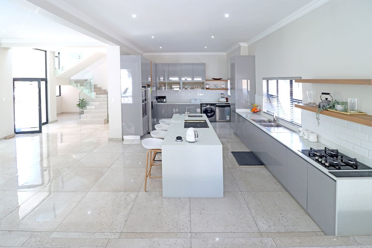 Modern open-plan kitchen featuring high-gloss grey cabinetry, stone countertops, a central island, and integrated gas hob, flowing into a bright staircase area.