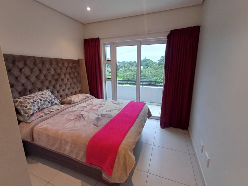 A well-lit bedroom featuring tiled flooring, recessed ceiling lights, and a sliding glass door providing access to a private balcony with scenic views.