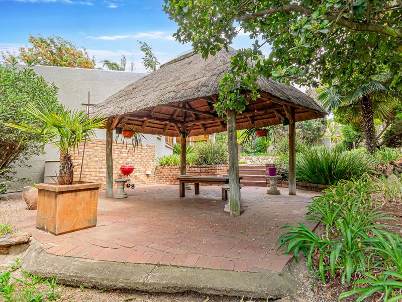 Outdoor entertainment area featuring a thatched gazebo structure set on a brick-paved patio, surrounded by established garden landscaping and brick retaining walls.
