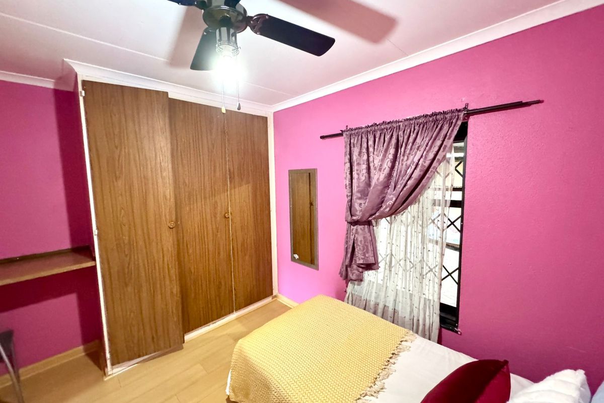A bedroom featuring built-in wooden wardrobes, a ceiling fan, and laminate flooring, finished with vibrant wall paint and security bars on the window.