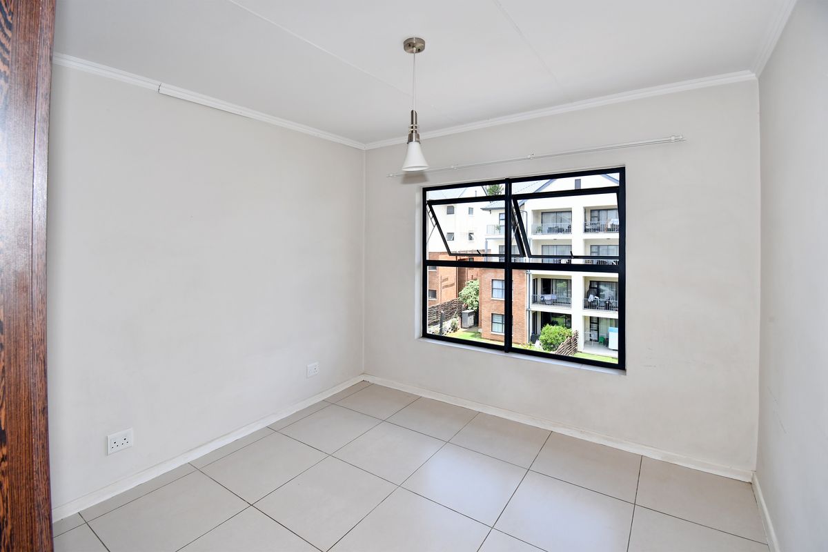 A bright, empty bedroom featuring neutral floor tiles, a modern pendant light, and a large window with black framing overlooking the apartment complex.
