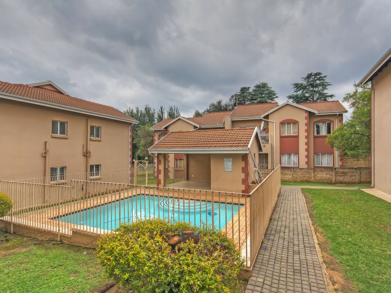 Communal pool area featuring a fenced swimming pool, paved pathways, and surrounding landscaped garden within an apartment complex.