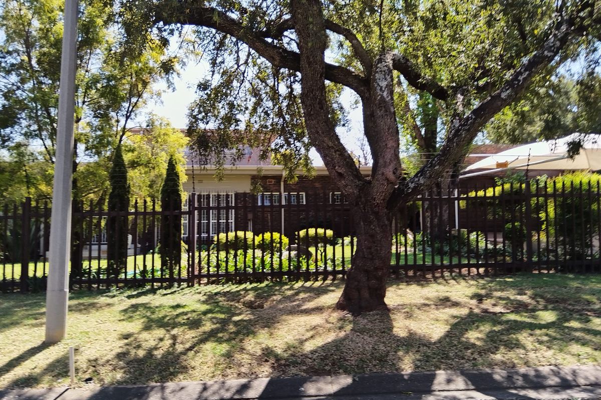 Street view of the property featuring a brick facade, mature trees, and secure metal perimeter fencing with electric security wires.