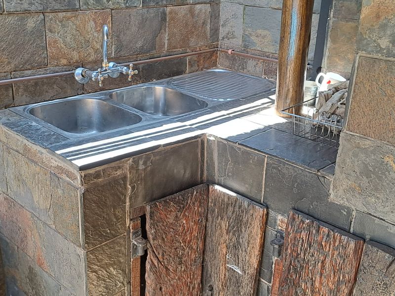 Outdoor scullery area featuring a stone-clad counter, double stainless steel sink, and rustic timber cabinetry with exposed copper plumbing.