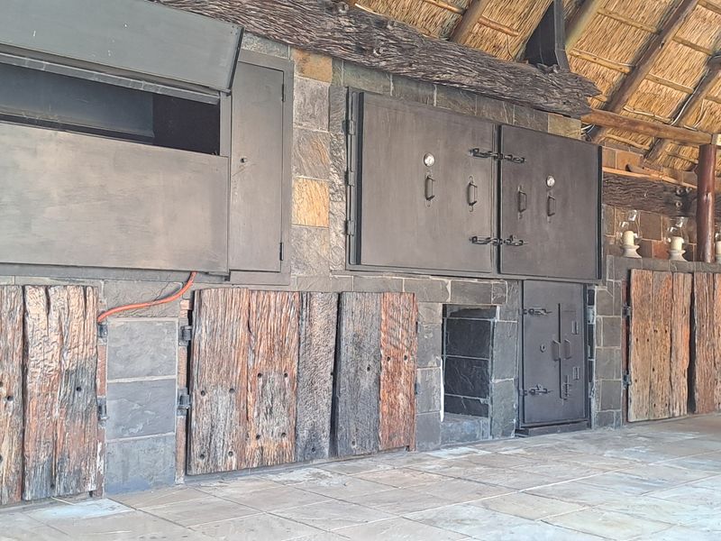 Covered outdoor entertainment area featuring a large built-in braai, rustic timber cabinetry, stone wall cladding, and a traditional thatched roof structure.