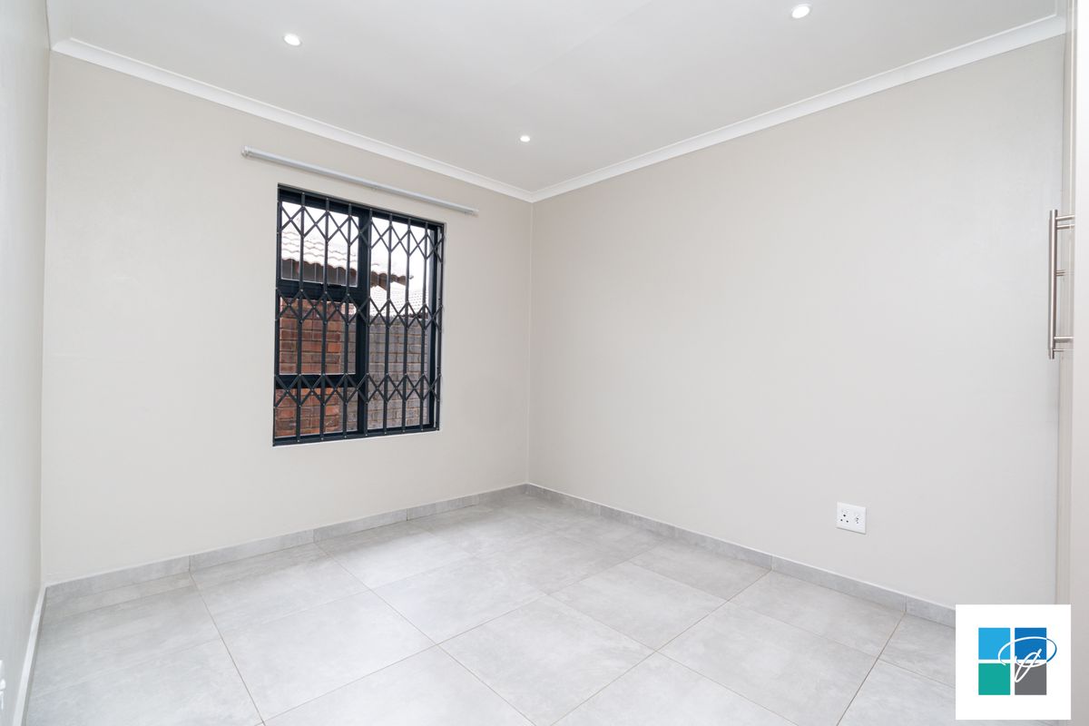 A bright, empty bedroom featuring neutral tiled flooring, recessed ceiling lighting, and a window fitted with a security gate.
