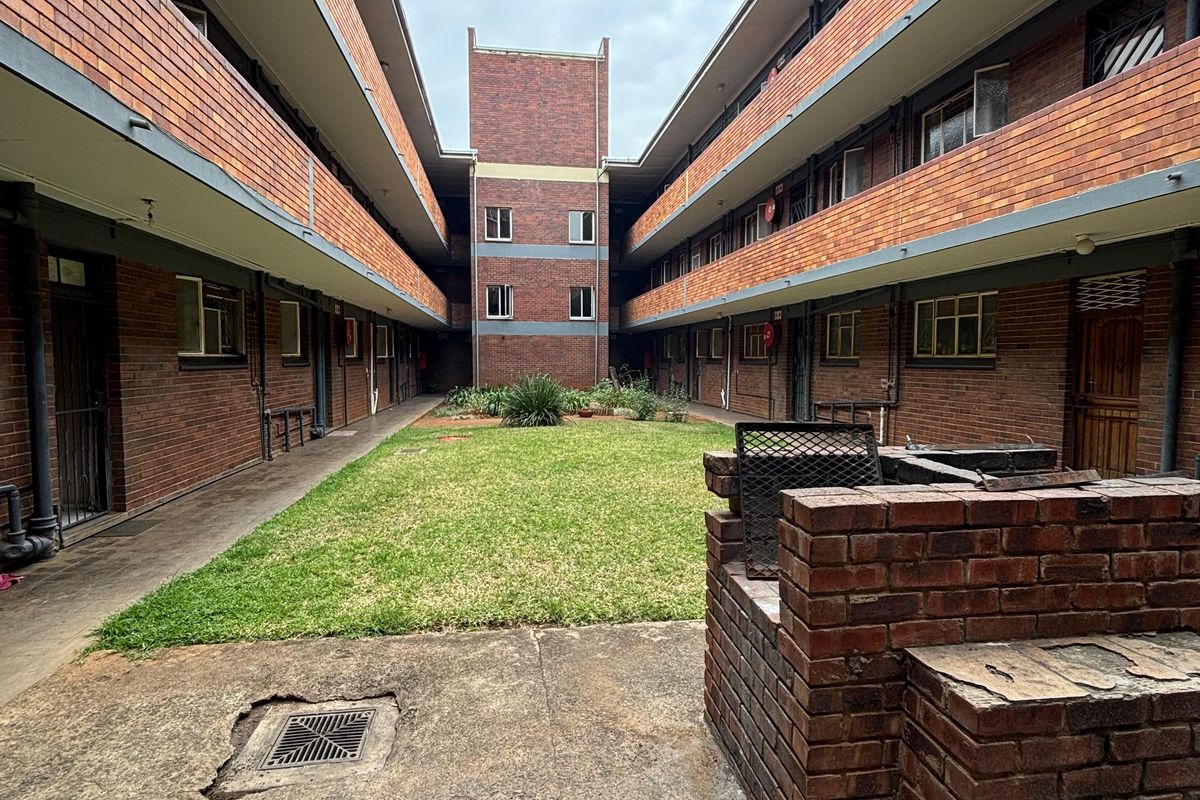 A communal courtyard area featuring a central lawn, brick apartment facades, and a built-in brick braai facility on a concrete patio.