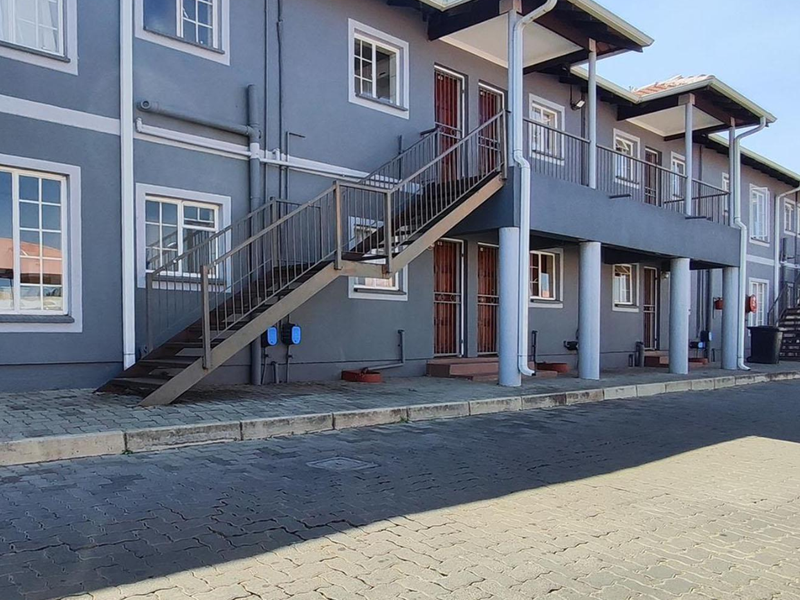 Exterior view of a multi-story apartment complex featuring grey facade, external metal staircases, and covered balcony walkways with paved ground access.