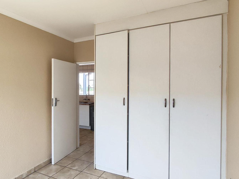 A clean, empty bedroom featuring neutral wall tones, tiled flooring, and ample built-in cupboard storage, with a view into the adjacent kitchen area.