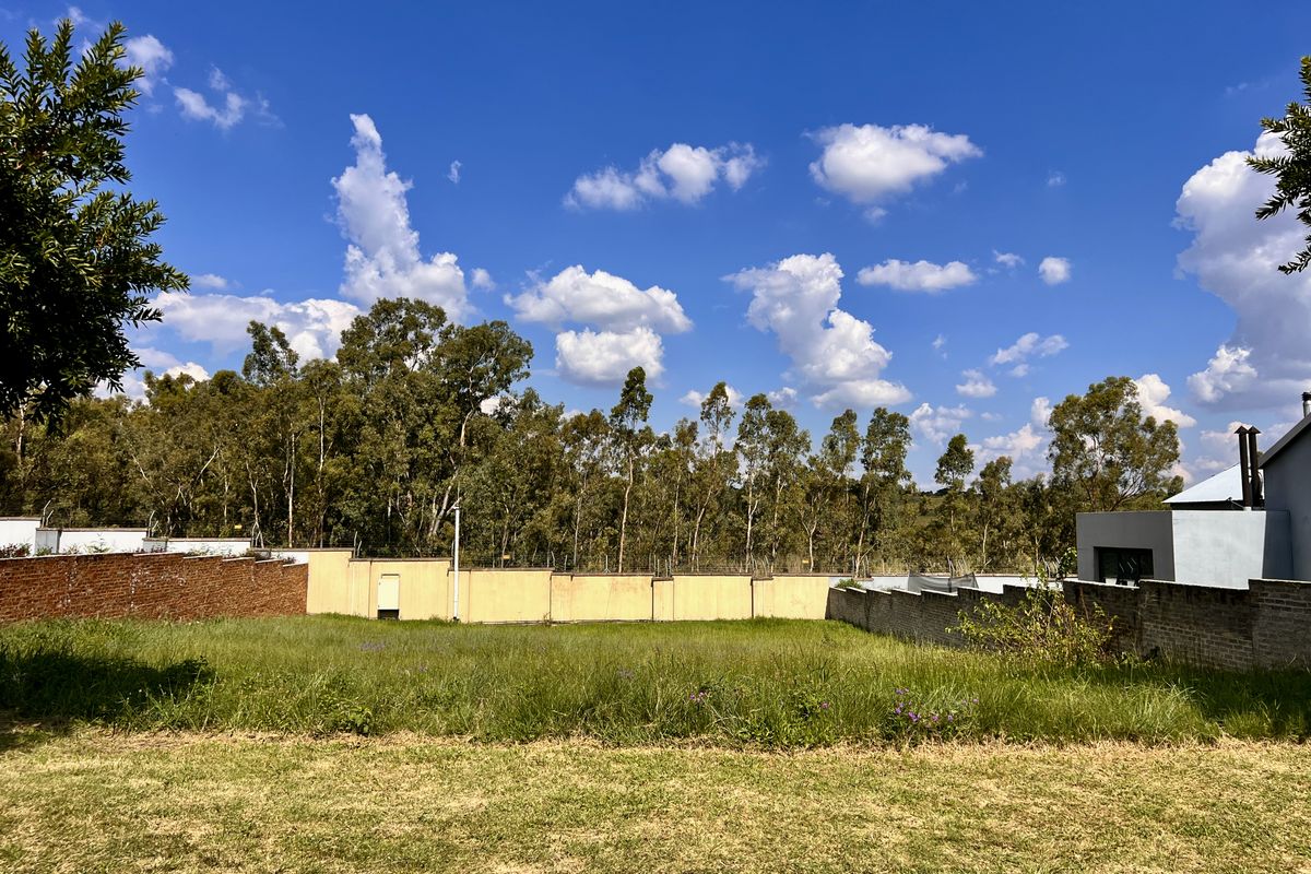 A vacant residential plot featuring a grass lawn, enclosed by a perimeter brick wall with integrated electric fencing and mature trees in the background.