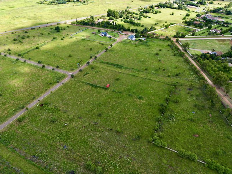 An aerial view of a large, undeveloped residential land plot featuring surrounding greenery, established road access, and perimeter fencing.