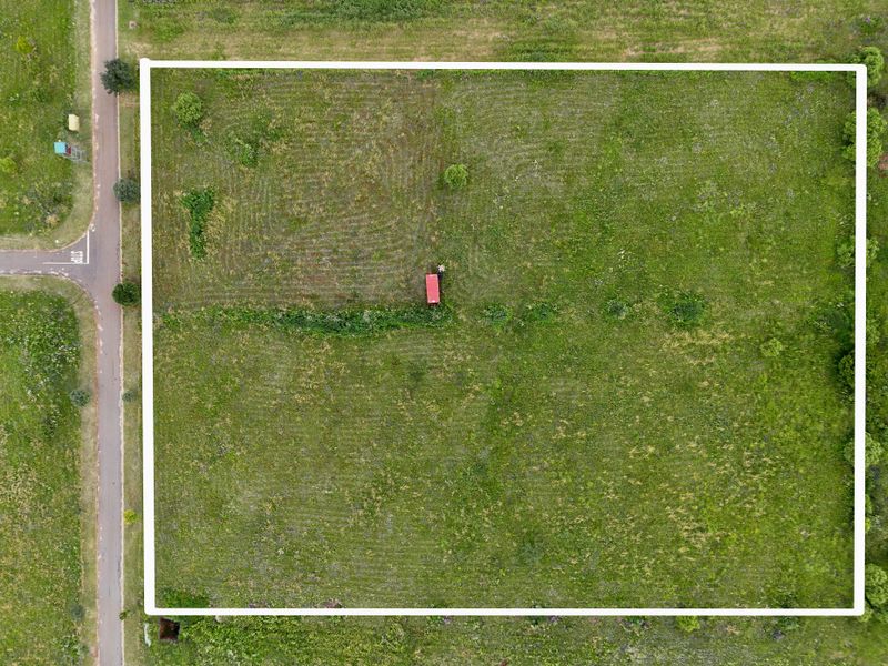 An aerial view of a vacant residential plot of land, featuring open grass coverage and direct access to the adjacent road.