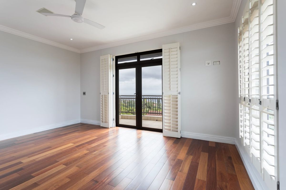 Bright and spacious bedroom featuring polished hardwood flooring, white plantation shutters, and direct access to a private balcony through double glass doors.