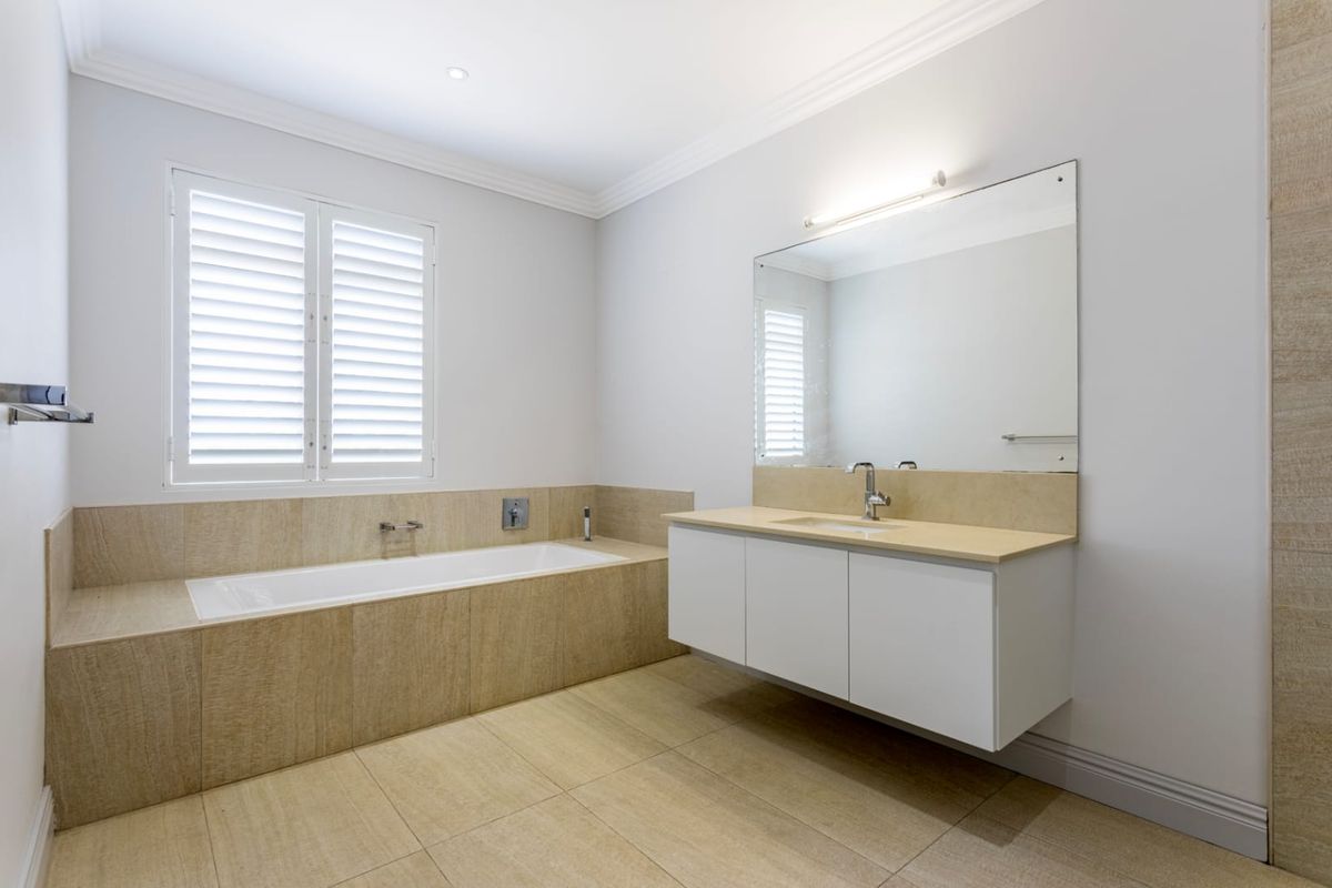 A modern, well-maintained bathroom featuring a floating vanity, large wall-mounted mirror, and a tiled built-in bathtub with plantation shutters.