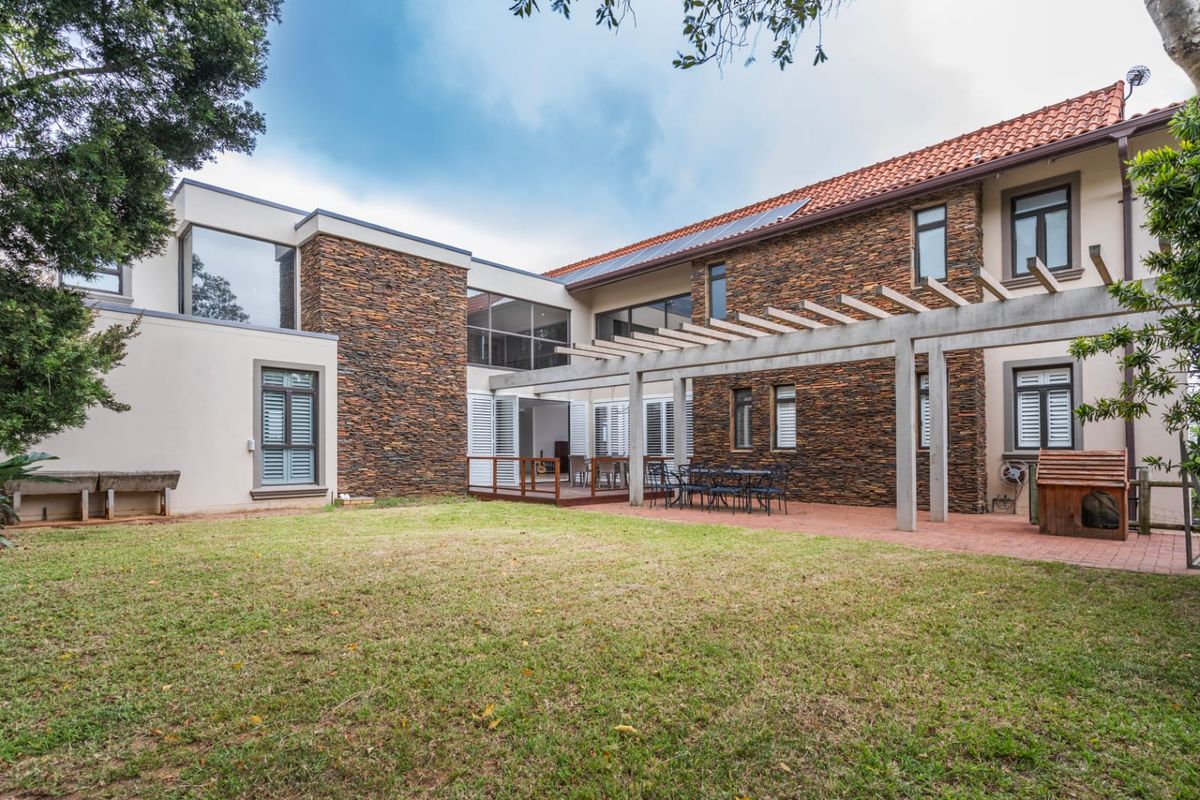Rear view of the property featuring a stone-clad facade, a paved patio area with a pergola, and a spacious lawn.