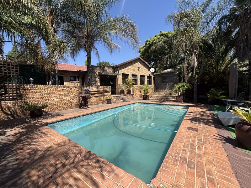 A well-maintained outdoor pool area featuring a rectangular swimming pool surrounded by brick paving and a brick retaining wall with mature landscaping.