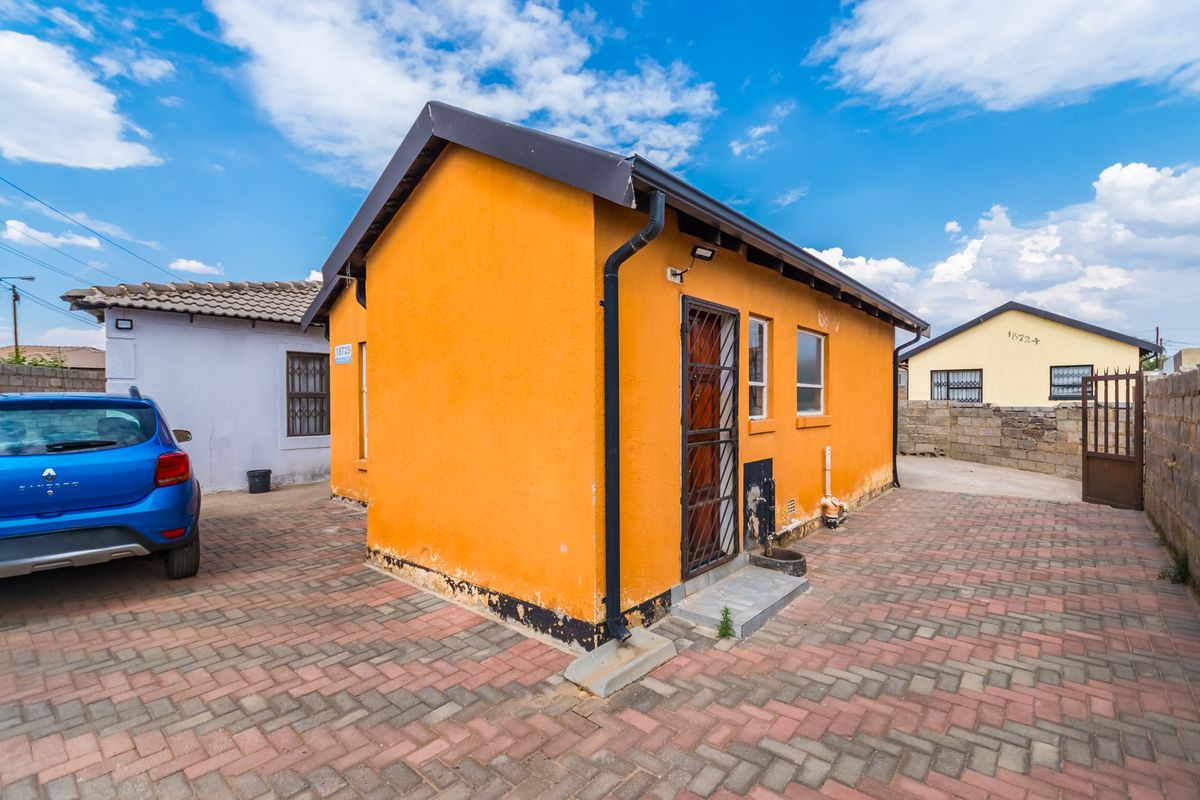 Rear exterior view of a detached house featuring a paved courtyard, security gate, and orange rendered walls with some visible wear at the base.