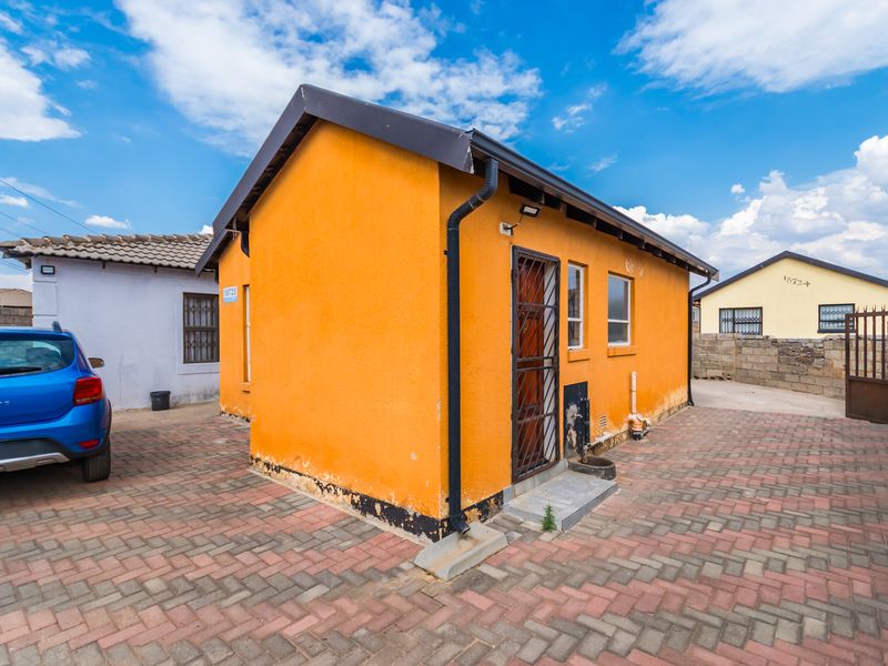 Rear exterior view of a detached house featuring a paved courtyard, security gate, and orange rendered walls with some visible wear at the base.