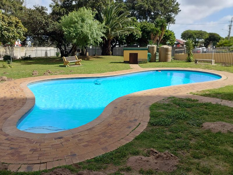 A communal outdoor pool area featuring a curved swimming pool with brick paving, surrounded by a landscaped garden and water storage tanks.