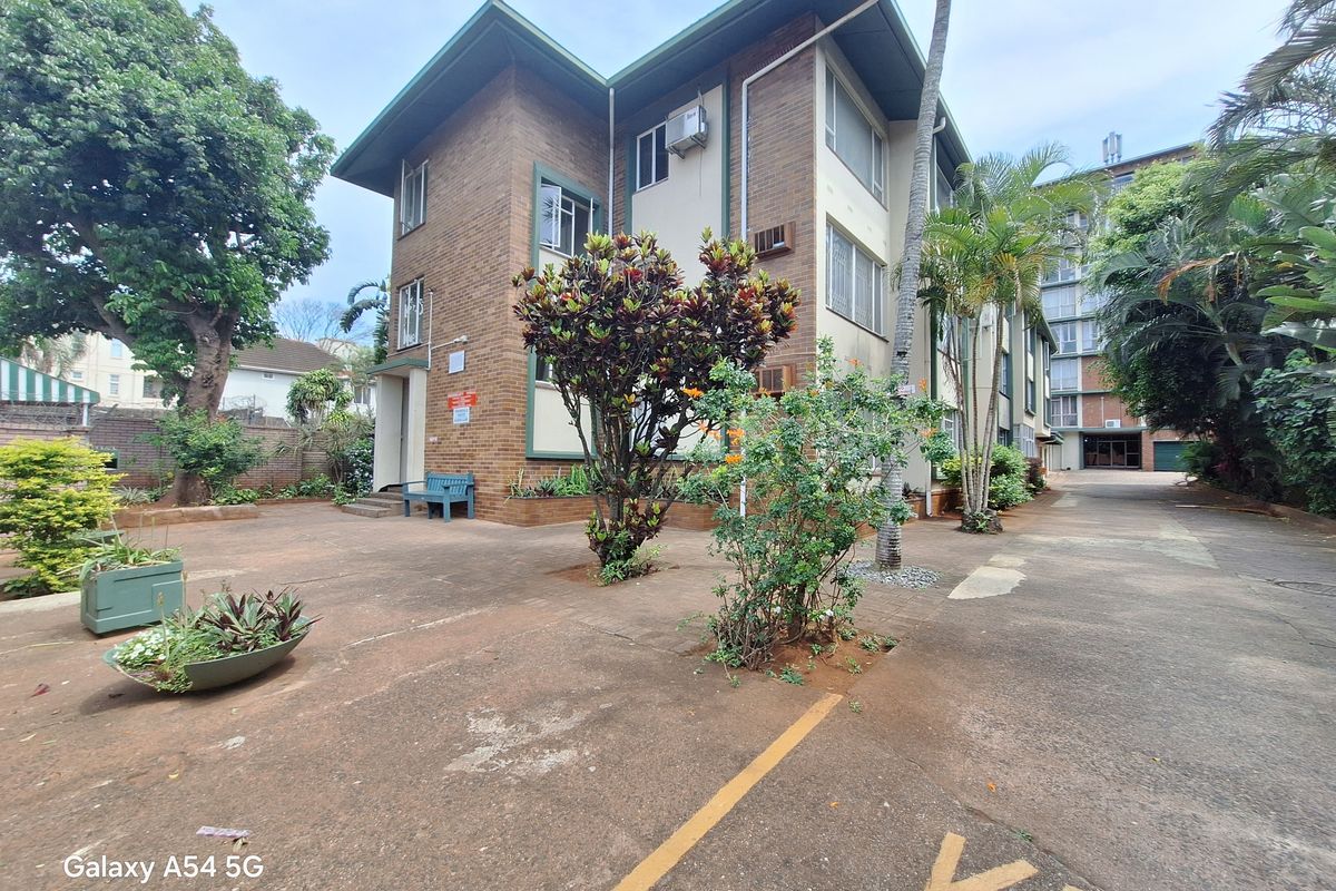 Exterior view of a multi-story brick apartment building featuring paved common areas, established landscaping, and dedicated parking spaces.