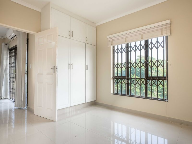 A bright, empty bedroom featuring built-in cupboards, tiled flooring, and a window fitted with a security gate.