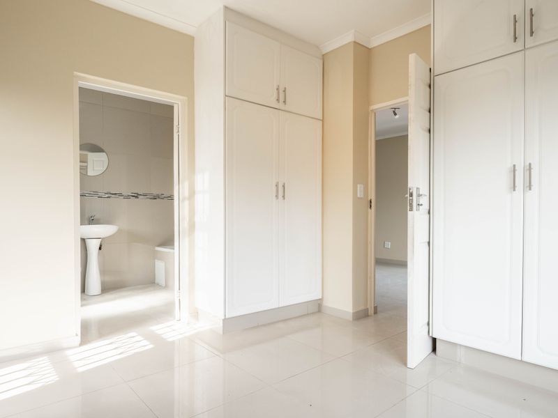 A bright, empty bedroom featuring built-in cupboards, tiled flooring, and an en-suite bathroom with a pedestal basin.