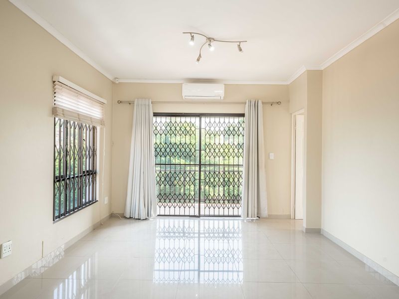 A bright, empty living room featuring tiled floors, a split-system air conditioner, and sliding glass doors with security gates leading to an outdoor area.
