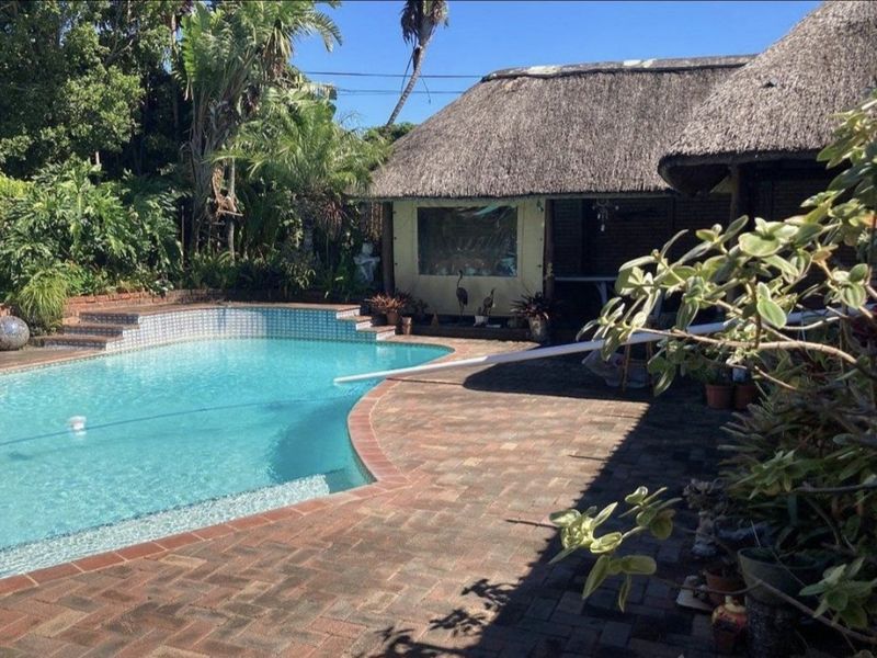 Outdoor pool area featuring a swimming pool with brick paving and a thatched roof structure in a landscaped garden setting.