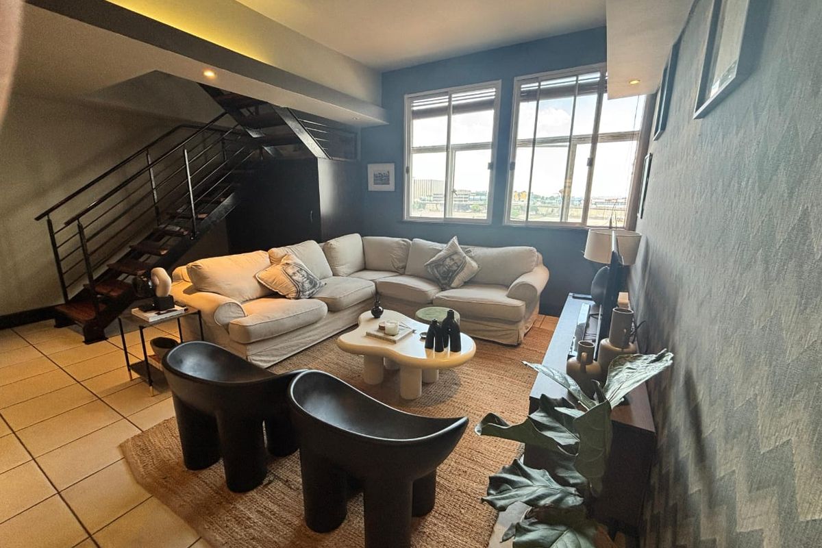 A spacious living room featuring a modern metal staircase, tiled flooring, and large windows that provide ample natural light.