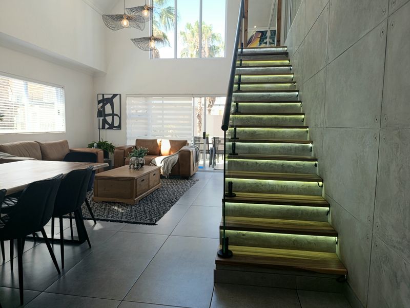 Spacious open-plan living area featuring a modern staircase with integrated LED lighting, concrete feature wall, and double-volume ceilings with large windows.