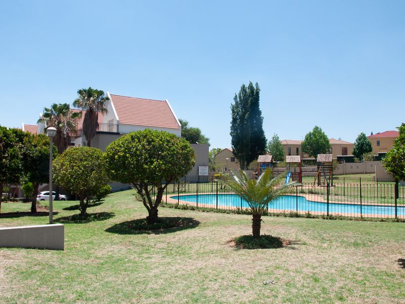 A well-maintained communal pool area featuring a swimming pool, metal fencing, and an adjacent children's play area set within a landscaped garden.