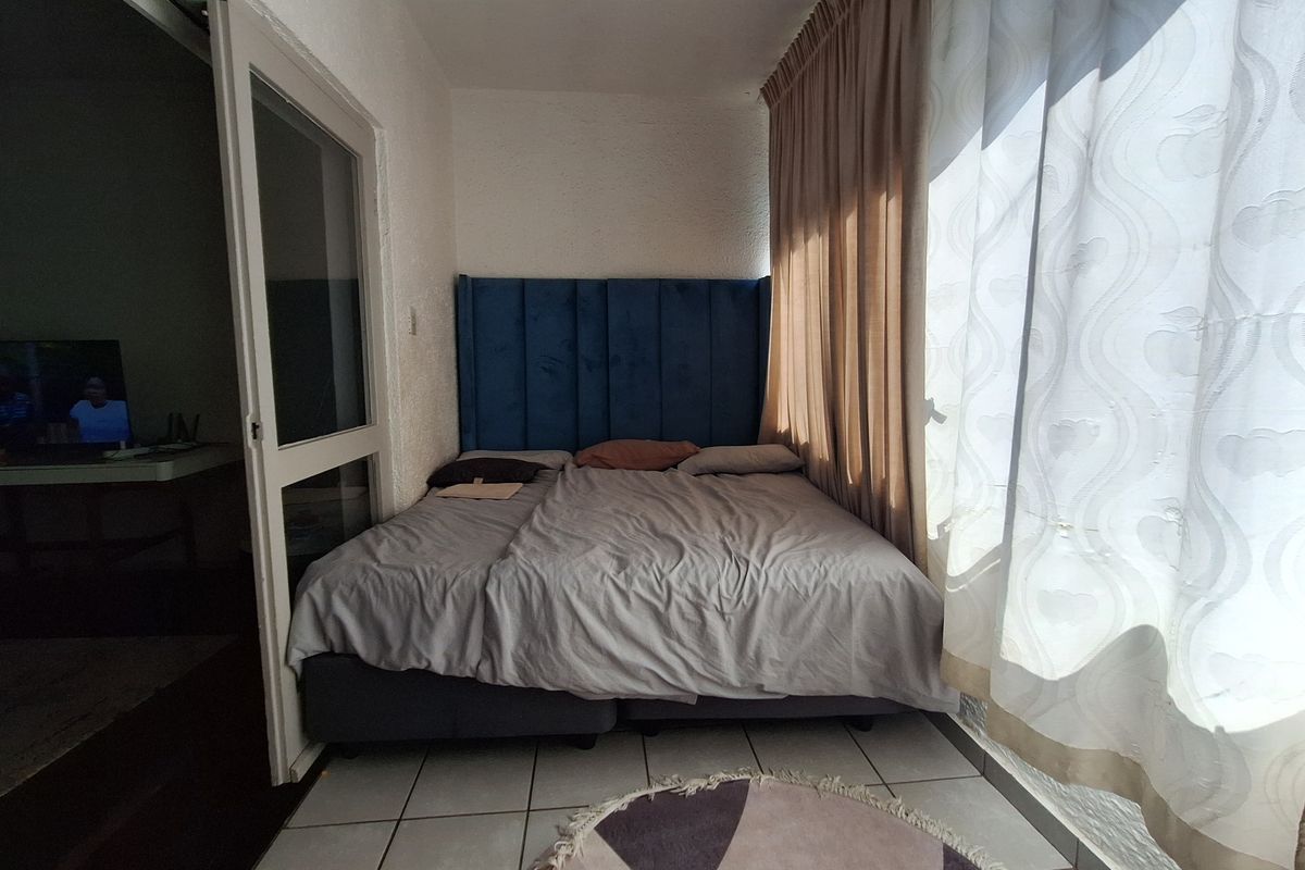 A compact bedroom featuring tiled flooring and a large window with curtains, providing natural light and access to an adjacent living area.