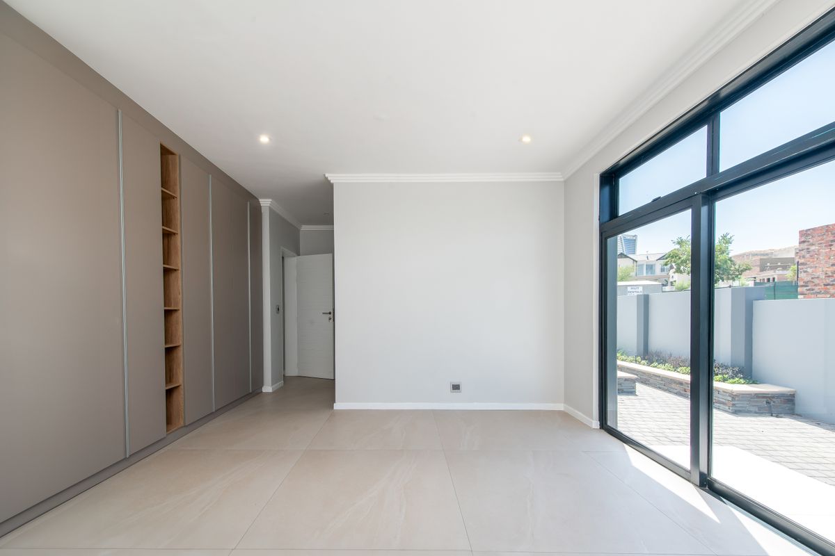 A bright, modern, and empty bedroom featuring extensive built-in cupboards, large sliding glass doors leading to the patio, and neutral tiled flooring.