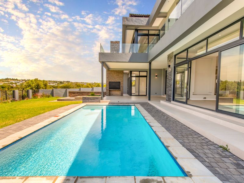 Modern outdoor entertainment area featuring a sparkling swimming pool, paved patio, built-in braai, and seamless indoor-outdoor flow through large sliding glass doors.